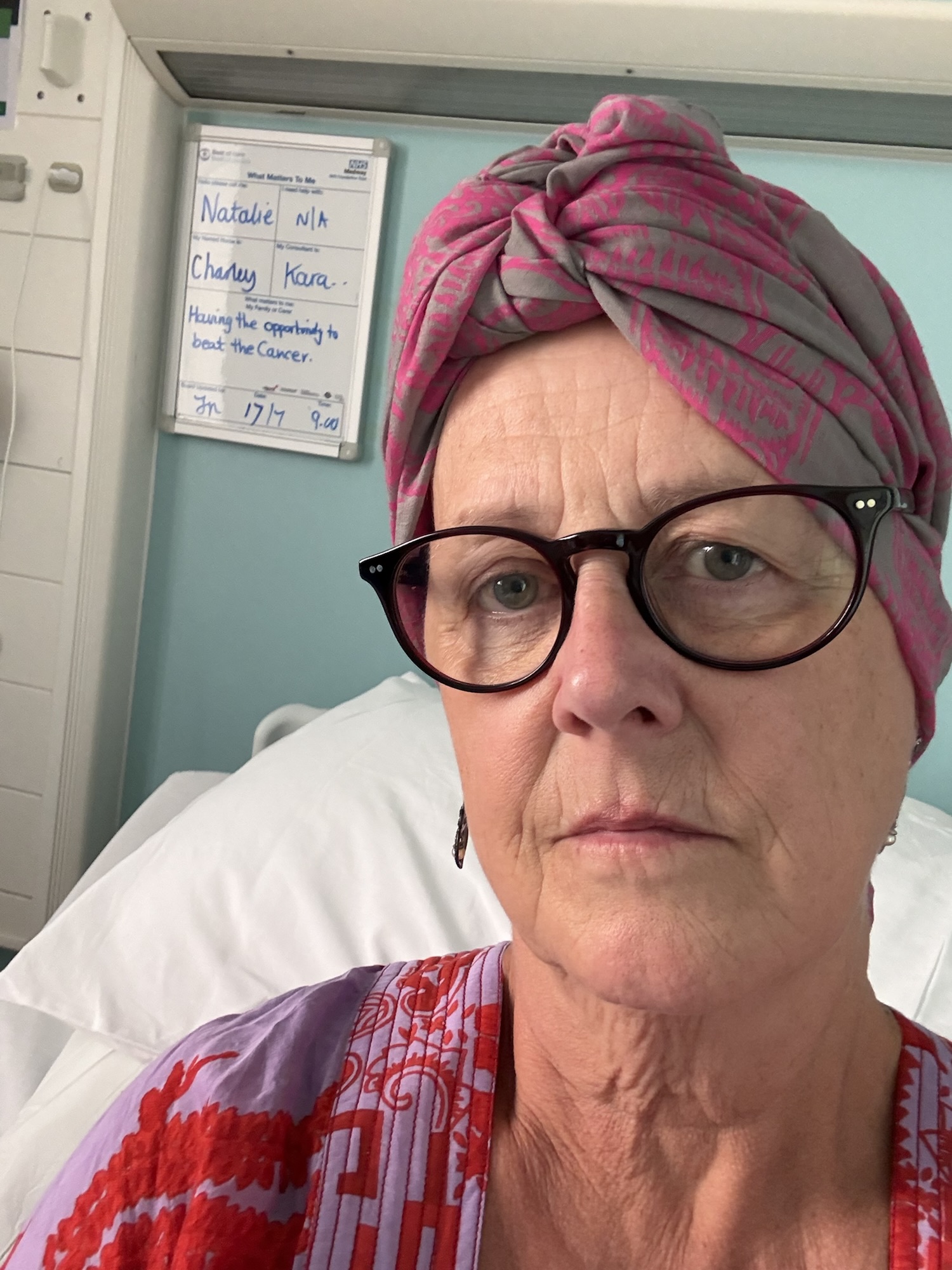 The author wearing glasses and a pink patterned headscarf in a hospital room with a whiteboard containing some general information and that what matters to her is "Having the opportunity to beat the Cancer"