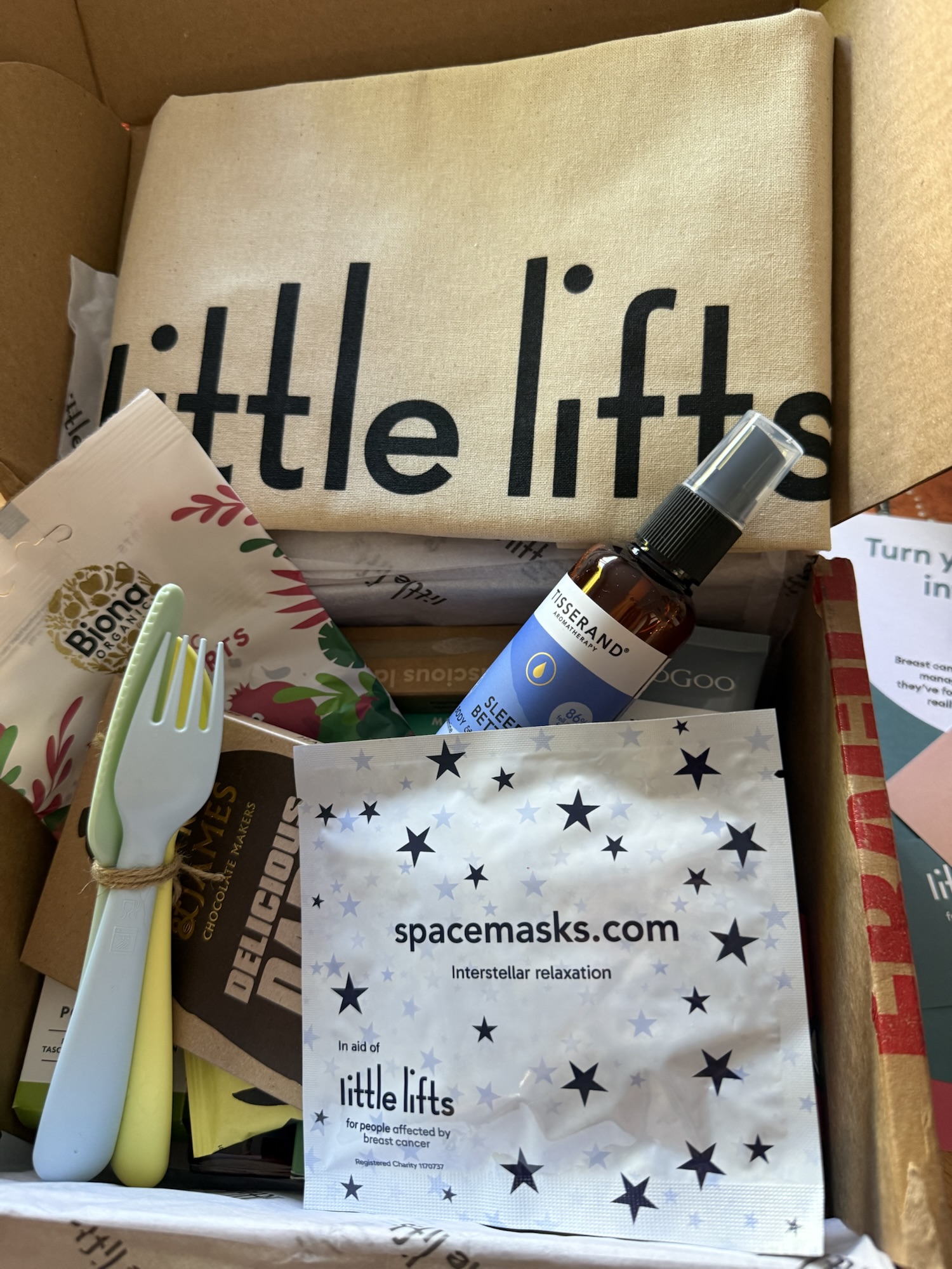 Box containing a folded beige fabric bag labeled little lifts, a packet labeled spacemasks.com interstellar relaxation, a Tisserand Sleep Better aromatherapy spray, Biona Organic utensils, and other small items