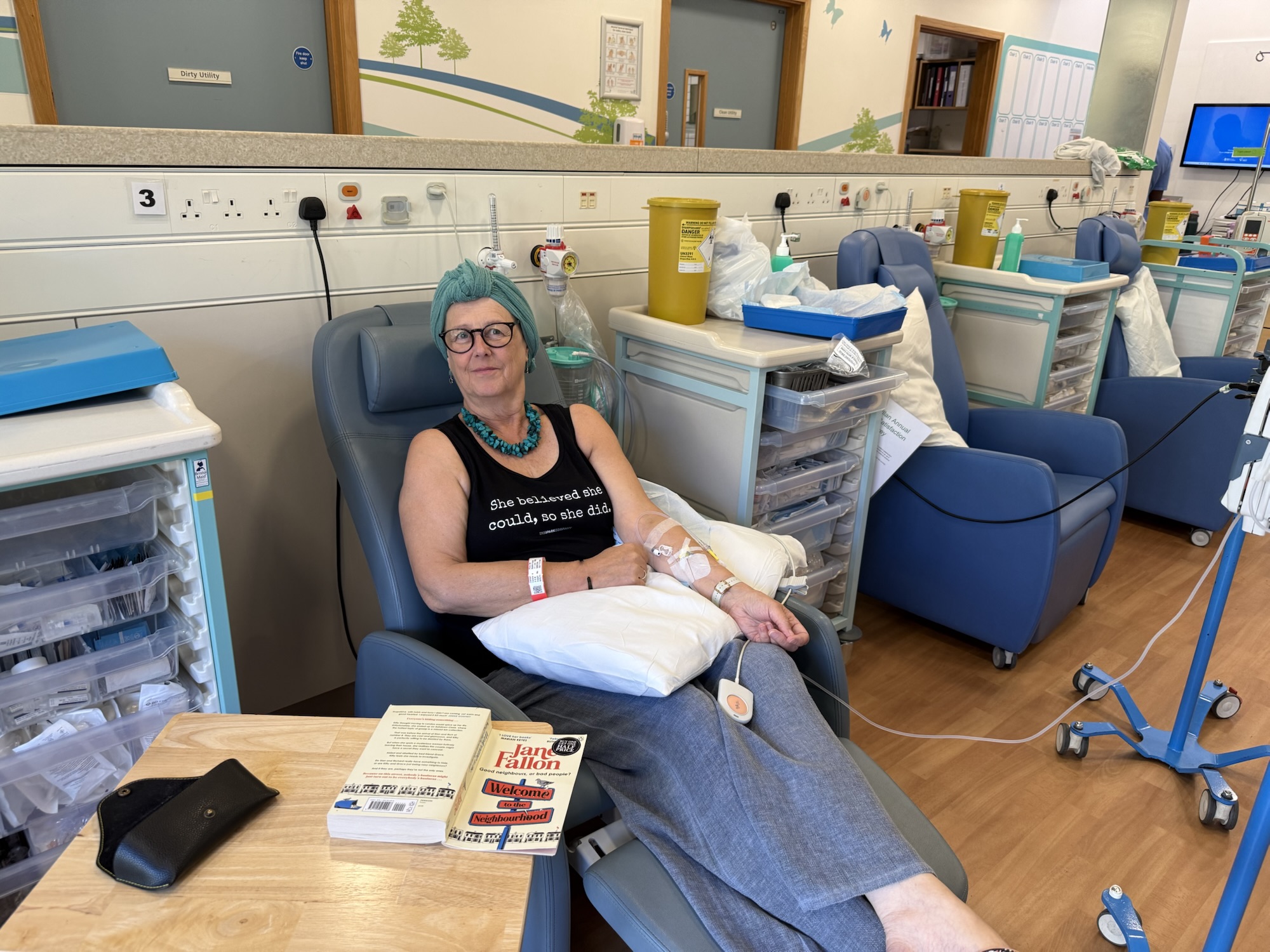 Treatment room Woman wearing a teal headscarf and necklace, and a vest that reads :"She believed she could, so she did." receiving medical treatment while seated in a recliner chair in a healthcare facility with medical equipment and supplies nearby