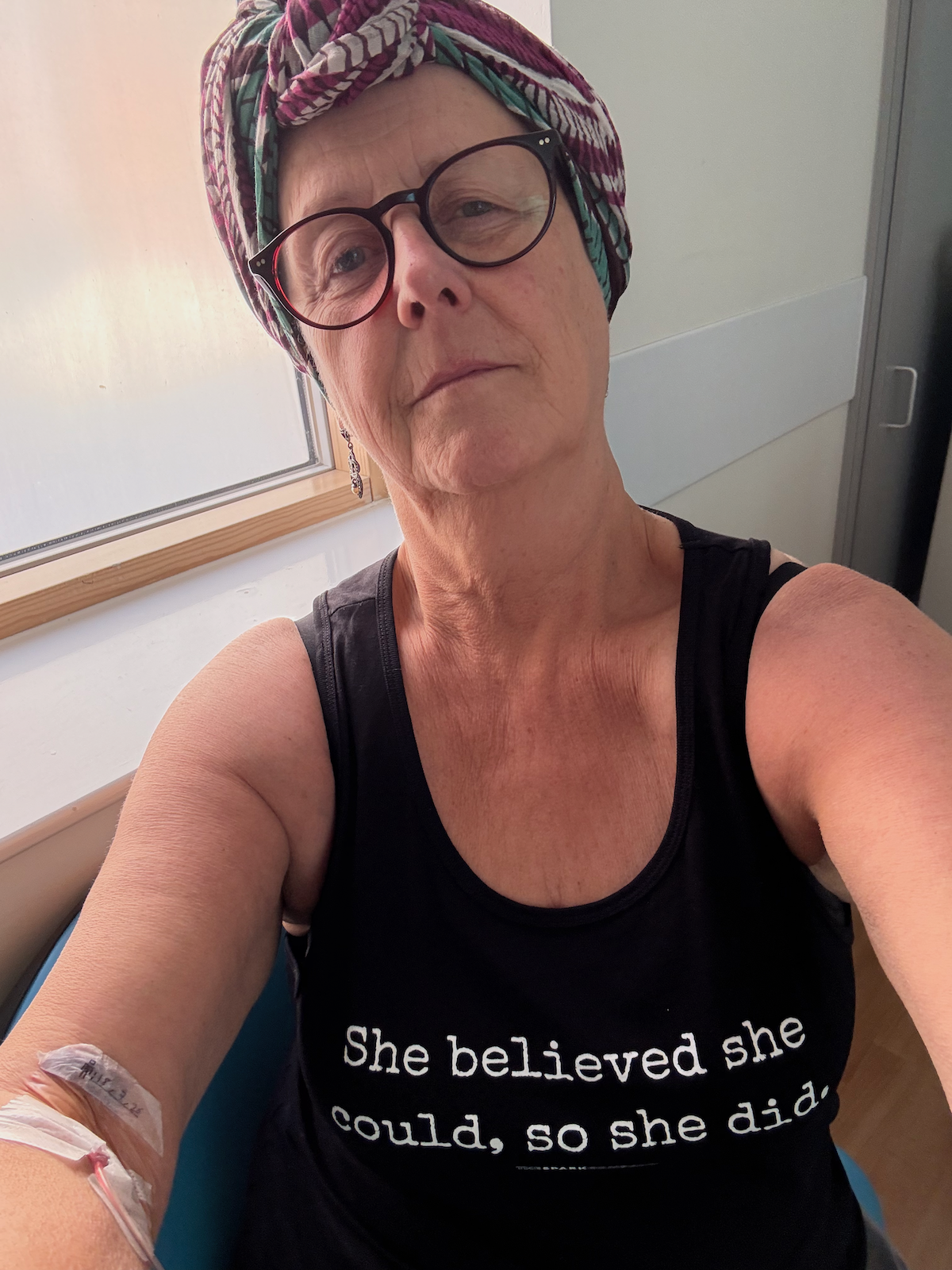 Woman wearing glasses and a colourful head wrap sitting by a window with a cannula in her arm and wearing a black tank top that reads 'She believed she could, so she did.'