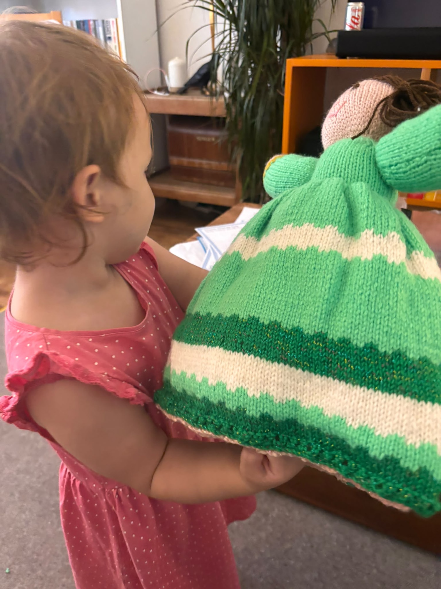 img_2171 A toddler wearing a pink sleeveless dress holding a knitted doll in green and white striped clothing