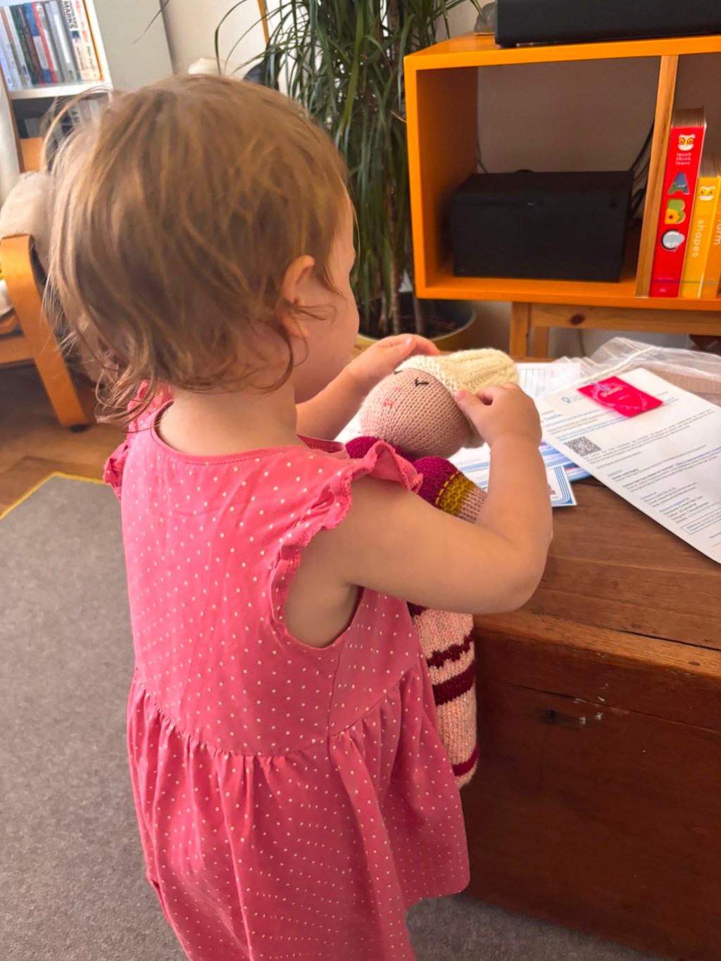 A child in a pink dress holding a knitted doll wearing a hat, standing near a wooden table with papers and a red packet on it