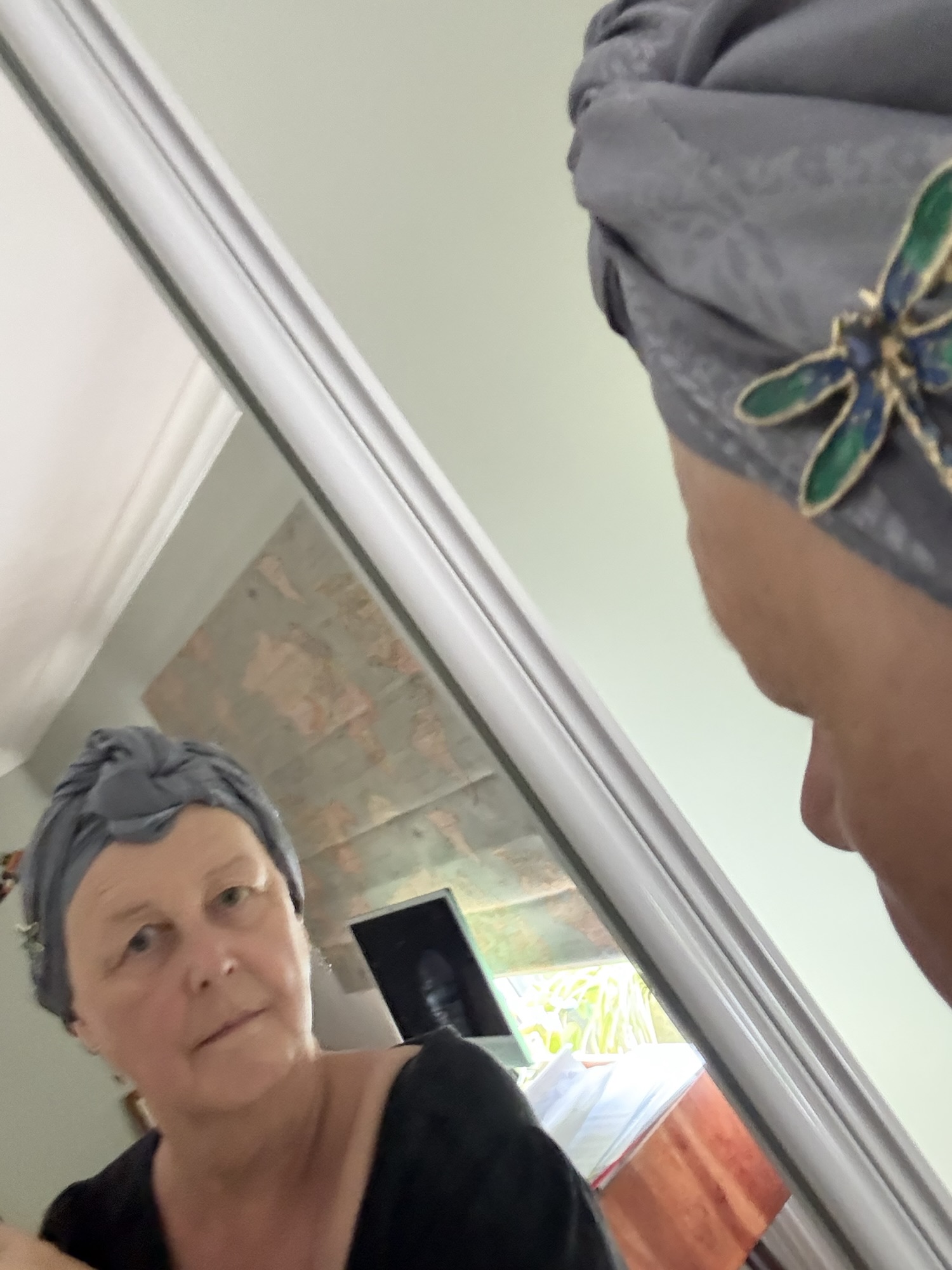A woman with a partially missing left eyebrow, wearing a silk turban with a dragonfly brooch, looks at herself in a mirror