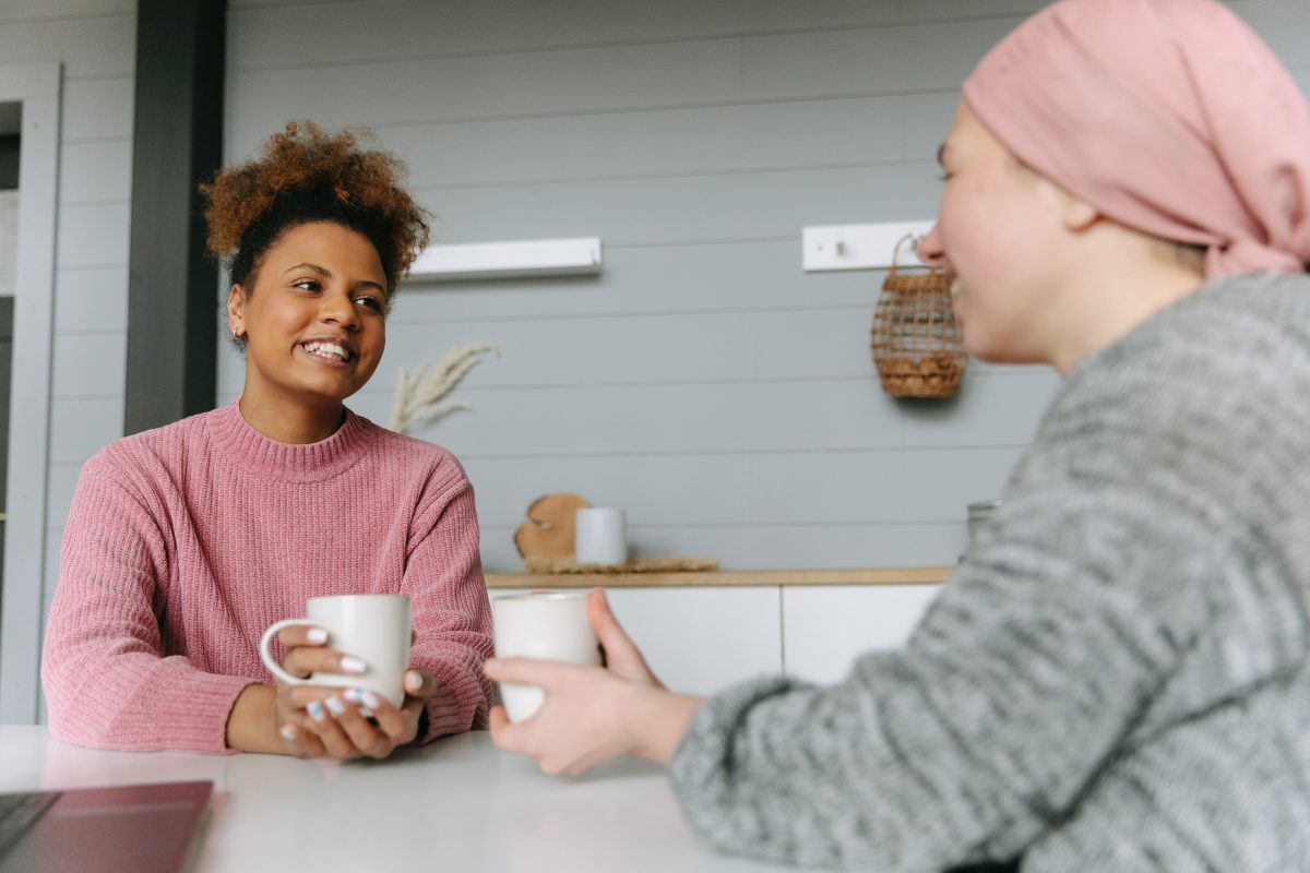 pexels-thirdman-7659464 Two women having a conversation, both are smiling and each holds a white mug, and one wears a headscarf.